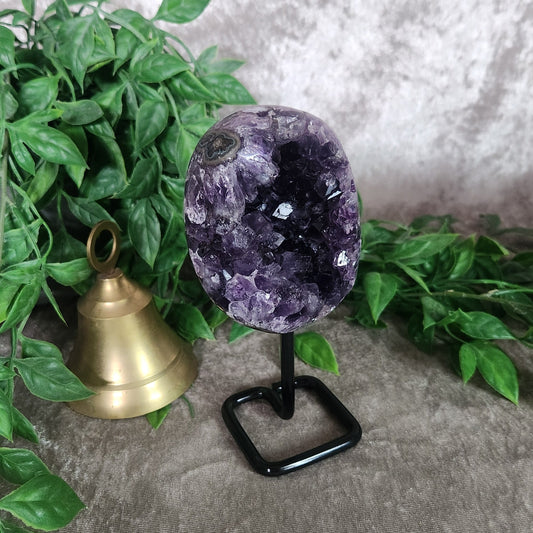 Amethyst geode on black metal stand with deep purple crystal points, styled with greenery and brass bell