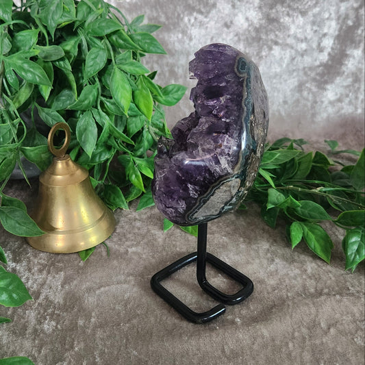 Amethyst portal geode on black metal stand with deep purple crystal points, displayed with greenery and brass bell