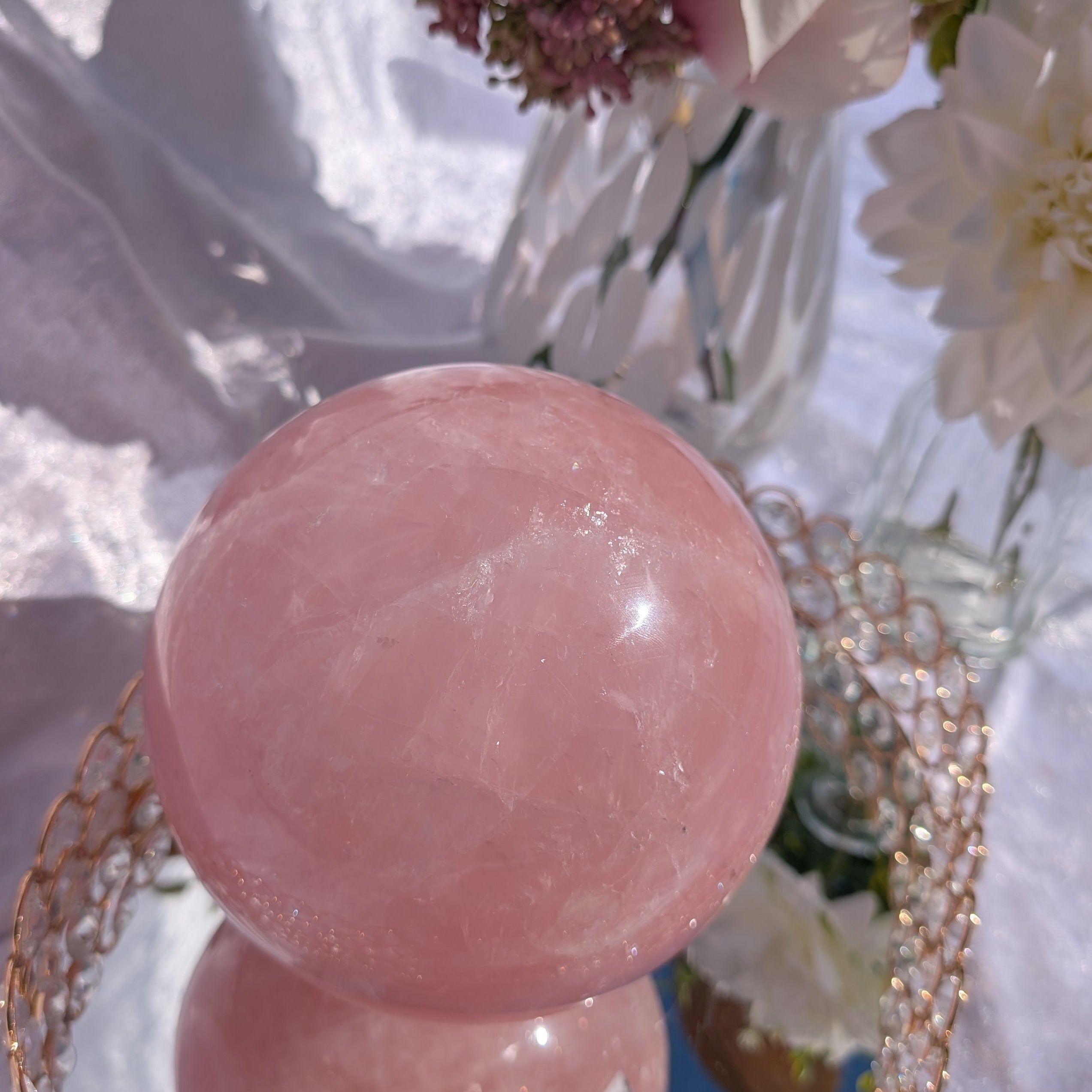 Rose Quartz Sphere – 4.38 kg polished sphere on mirrored tray surrounded by pink flowers and soft white fabric backdrop.