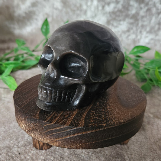 Silver sheen obsidian skull 446g displayed on wooden circular stand with greenery in background and soft velvet fabric