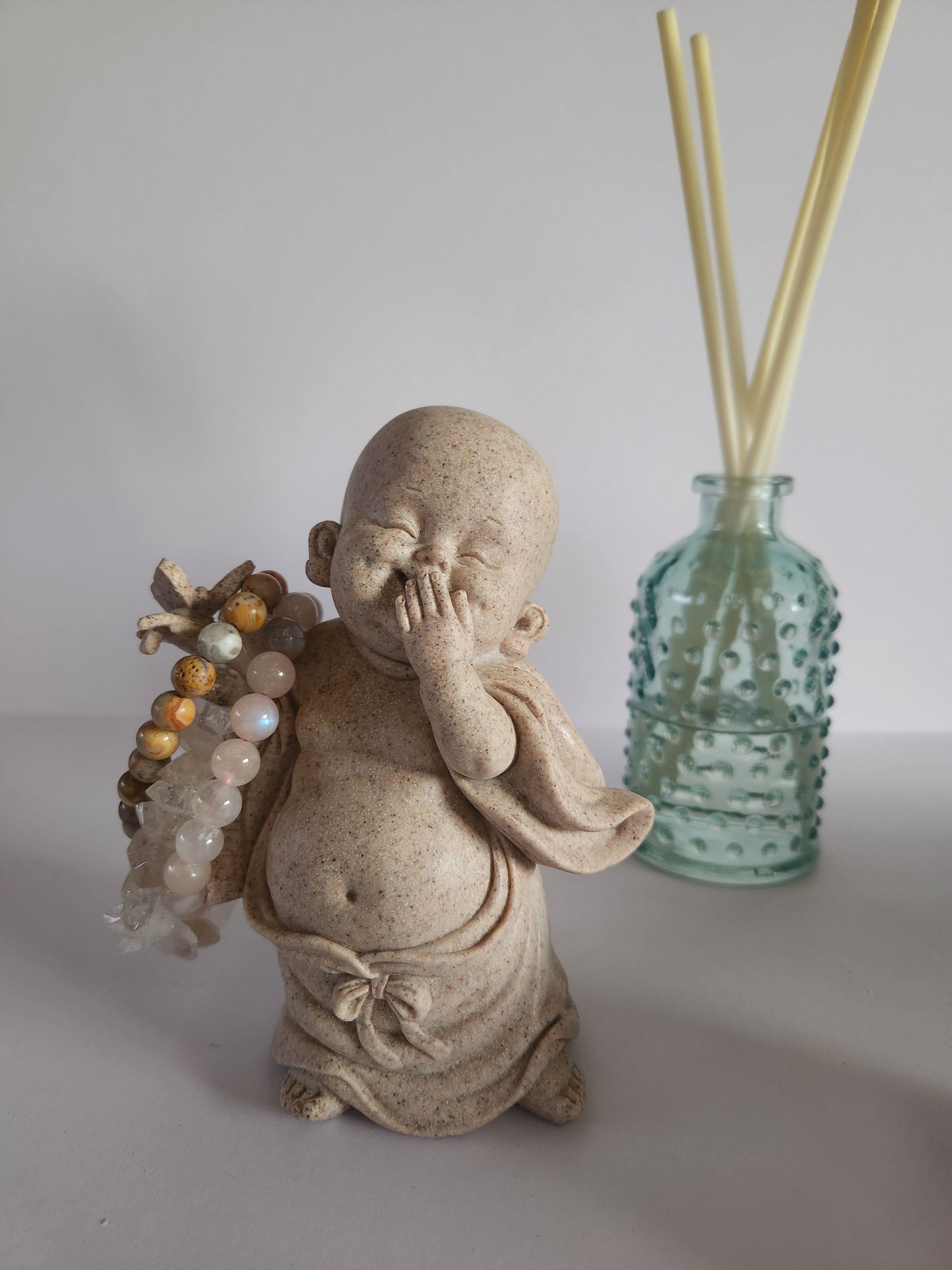 Smiling Buddha statue holding crystal bead bracelets beside glass diffuser. Spiritual décor with calming energy.